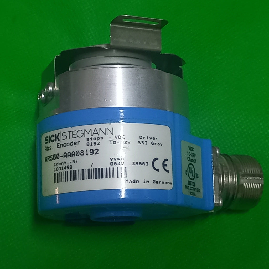A SICK STIGMANN industrial electronic component with a blue and silver body, featuring a white label with a barcode and technical specifications, photographed on a bright green textured background.