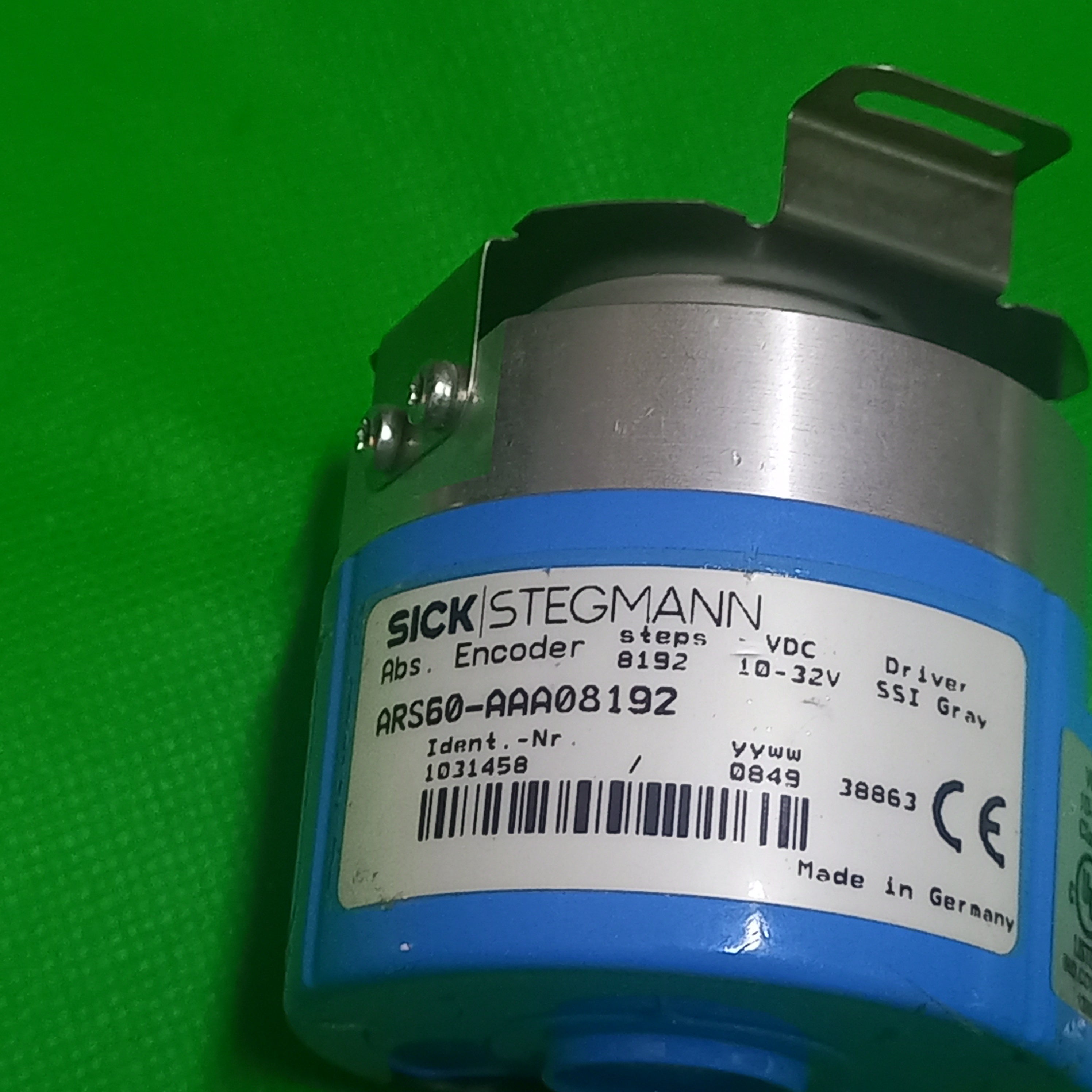 A Sick Stegmann rotary encoder module with a blue plastic housing, silver metal core, and a white product label, photographed on a bright green background.